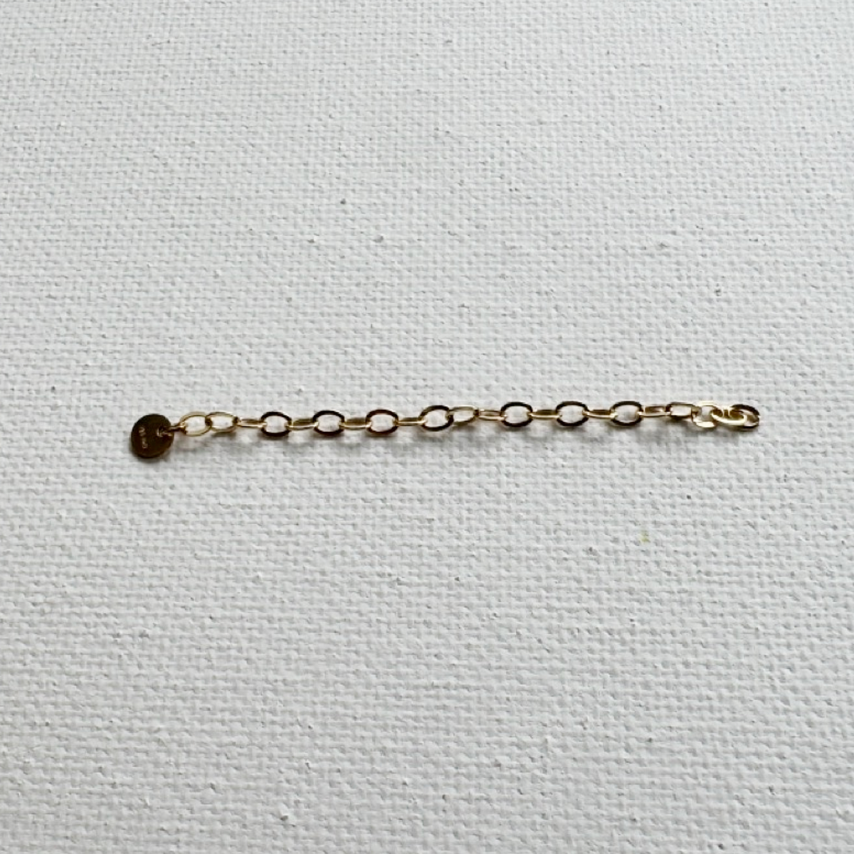 A delicate gold chain extender with a small round gold disc charm, laid horizontally on a textured white canvas background.