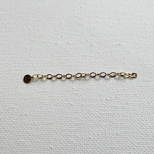A delicate gold chain extender with a small round gold disc charm, laid horizontally on a textured white canvas background.