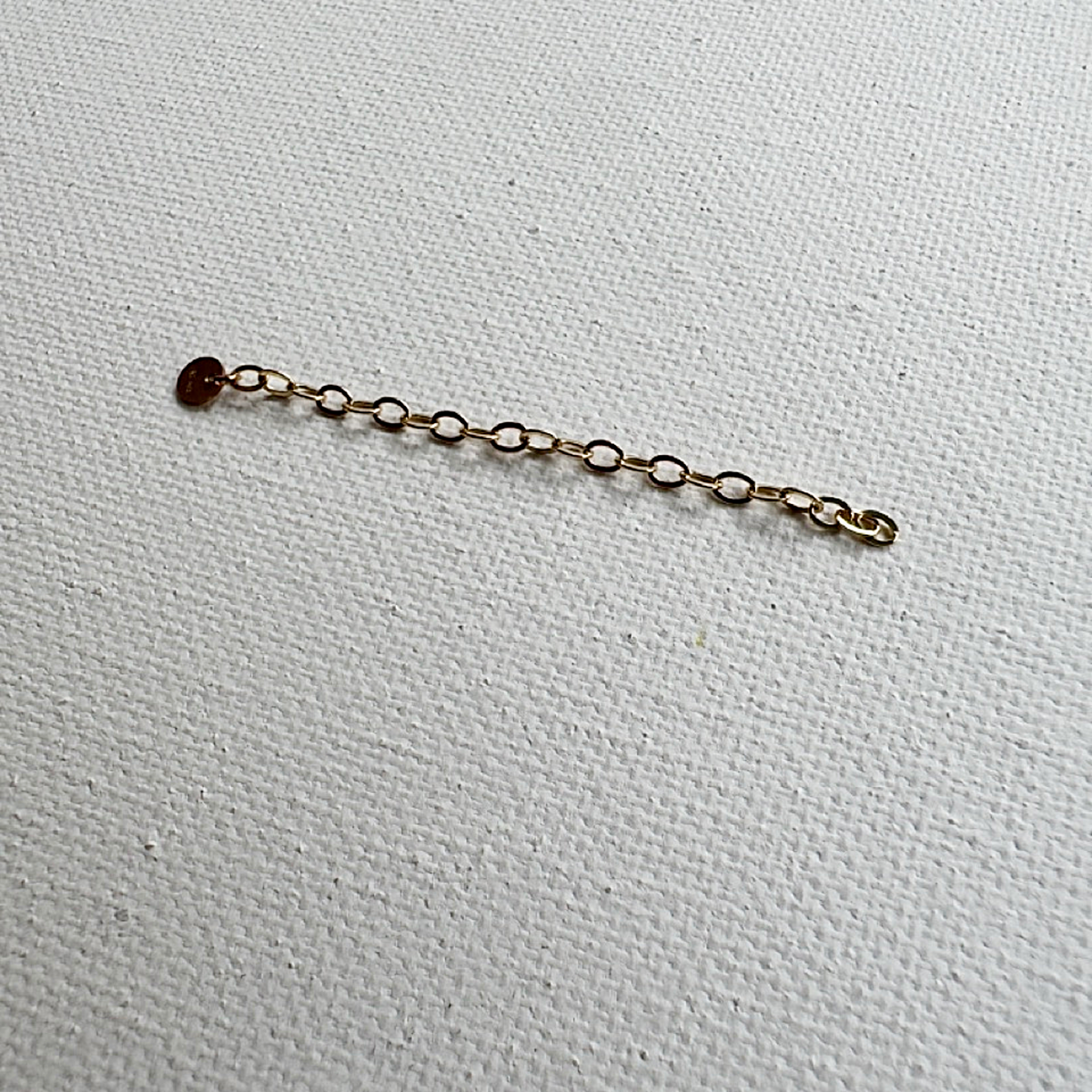 A delicate gold chain extender with a small round  pendant, laid flat on a textured white fabric background.
