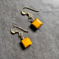 A pair of handmade dangle earrings featuring square yellow-gold tiger's eye stones on antiqued gold fishhook wires, displayed on a textured gray background.