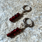 A pair of silver hoop earrings with dangling clusters of faceted dark red beads, displayed on a textured gray stone surface.