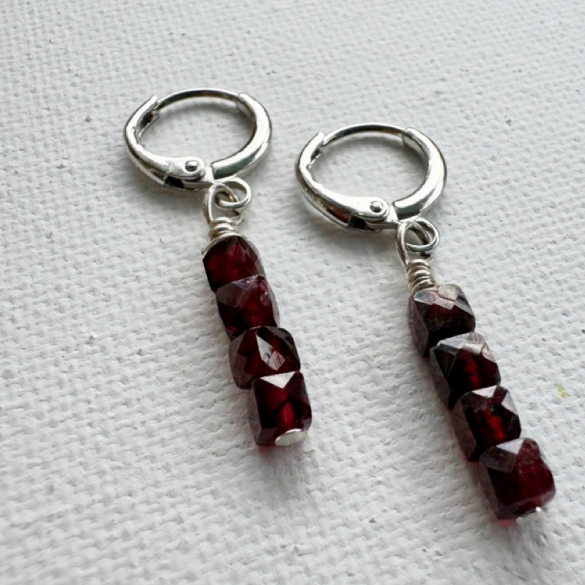 A pair of silver hoop earrings with faceted dark red beads dangling from silver wire loops, displayed on a textured white fabric background.