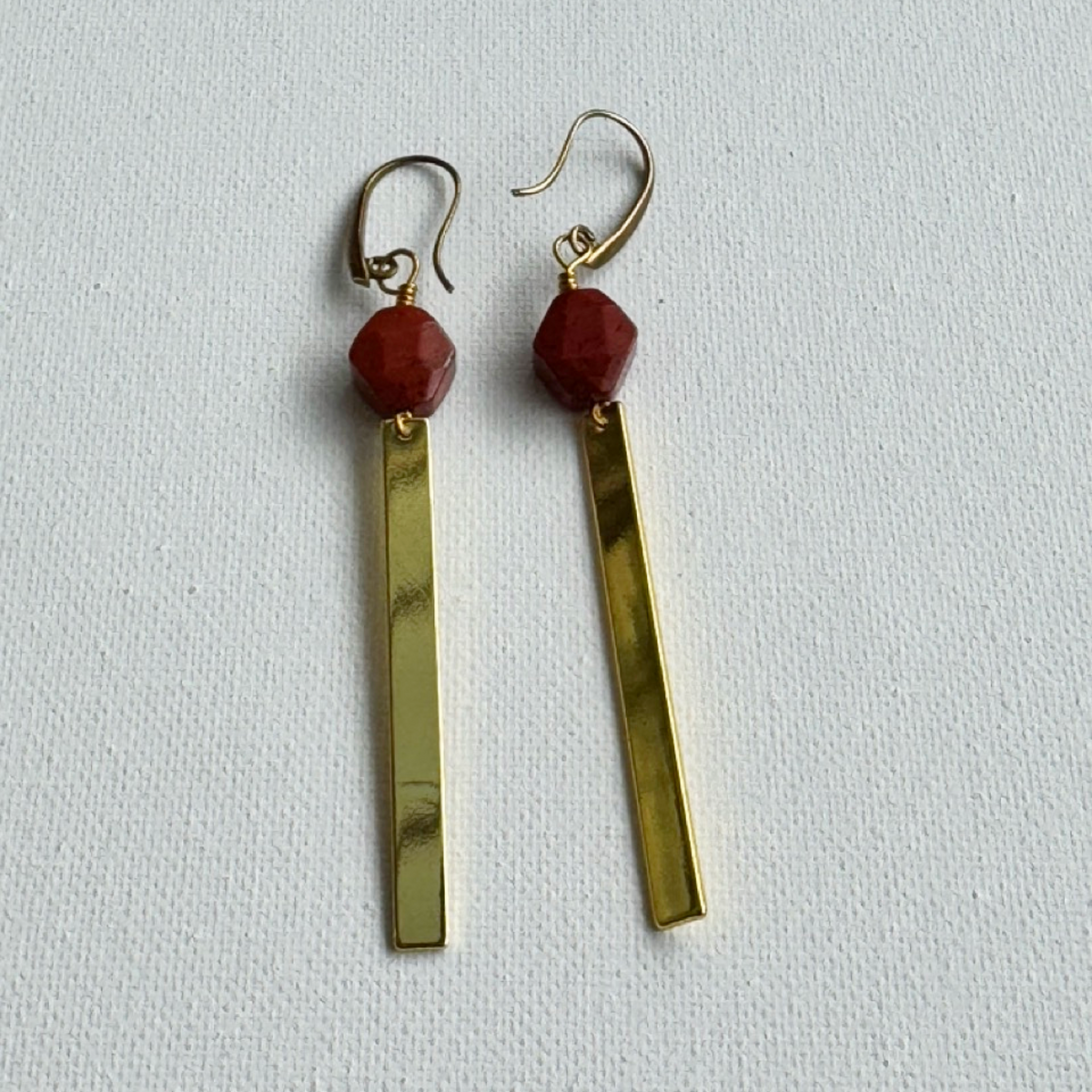 A pair of gold-toned dangle earrings with faceted red beads and long rectangular gold bars, displayed on a textured white background.