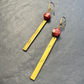 A pair of gold-toned dangle earrings with faceted red beads and long rectangular gold bars, displayed on a textured gray surface.
