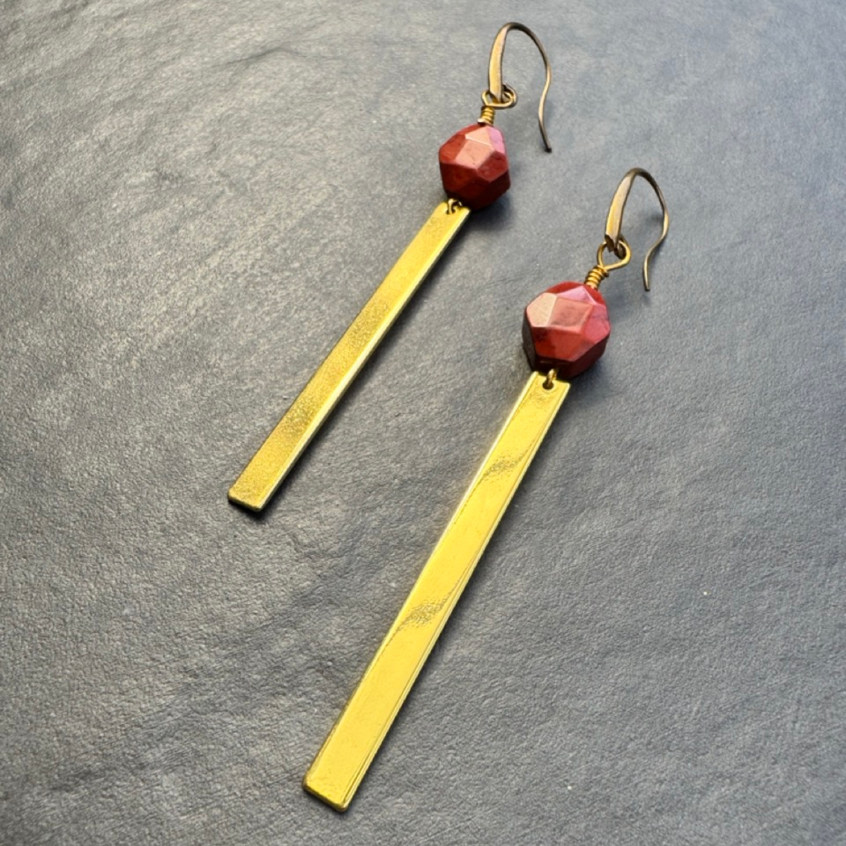 A pair of gold-toned dangle earrings with faceted red beads and long rectangular gold bars, displayed on a textured gray surface.