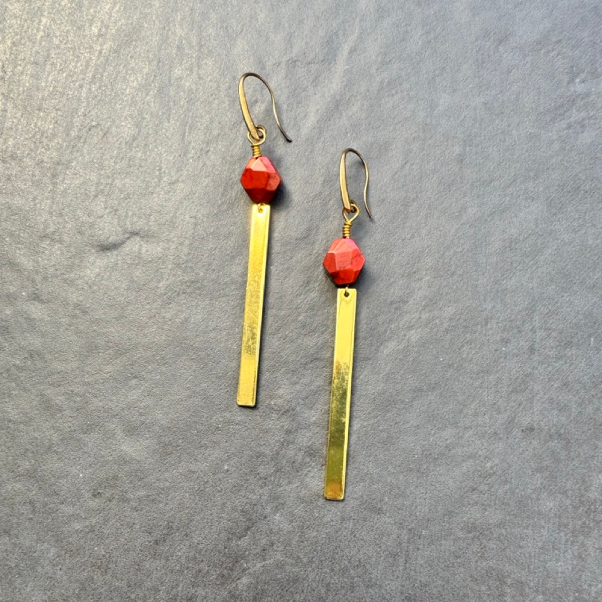 A pair of gold-toned dangle earrings with faceted red beads and long rectangular gold bars, displayed on a textured gray background.