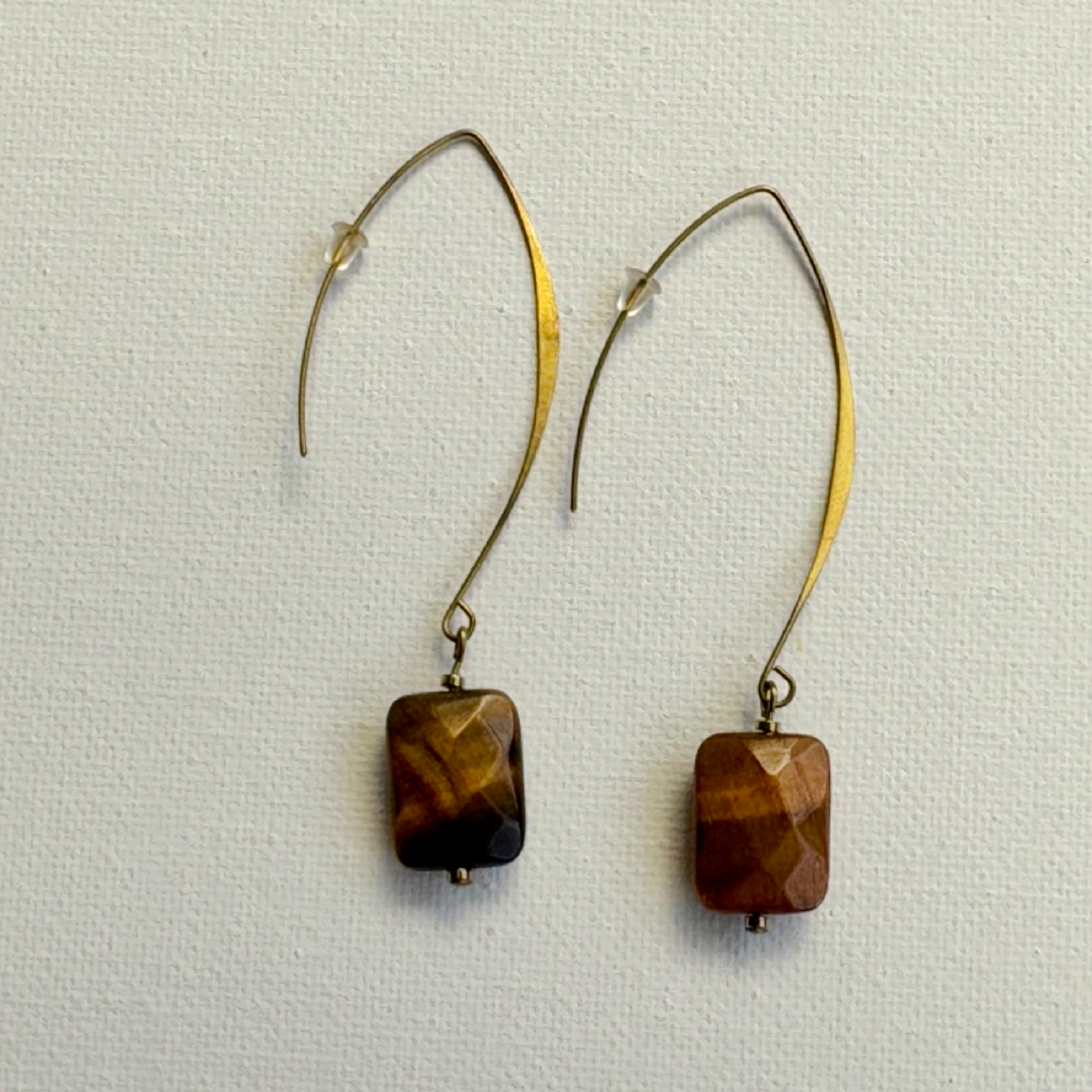A pair of dangling earrings with curved gold-tone hooks and faceted brown tiger's eye beads, displayed on a textured off-white background.