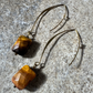 A pair of drop earrings with faceted tiger's eye beads and long curved gold-tone wire hooks, displayed on a textured gray stone surface.