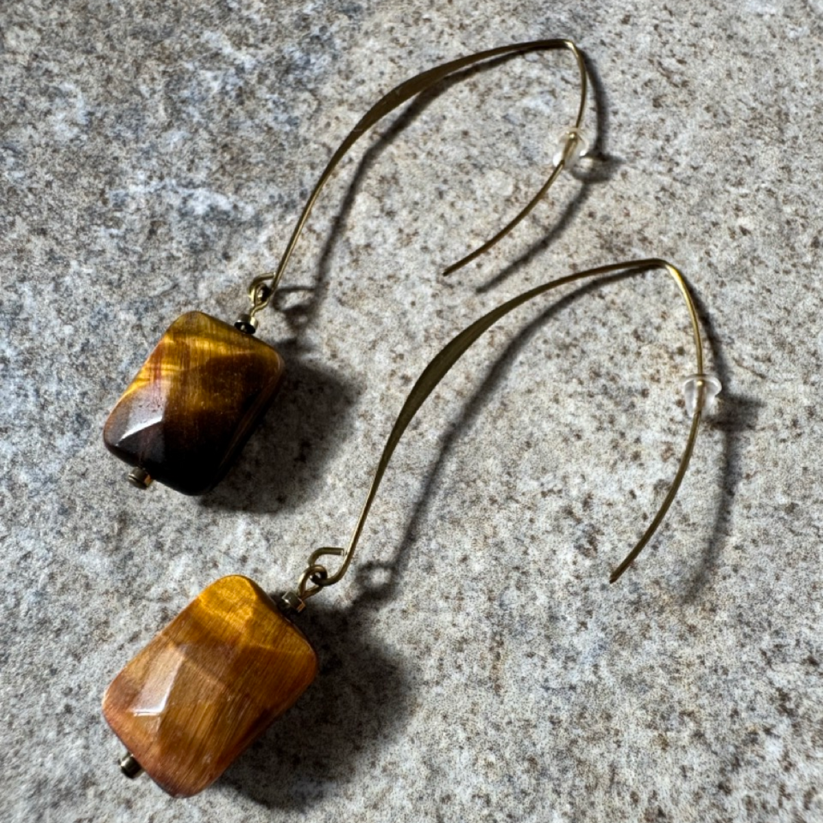 A pair of drop earrings with faceted tiger's eye beads and long curved gold-tone wire hooks, displayed on a textured gray stone surface.