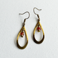 A pair of teardrop-shaped dangling earrings with an antique brass finish, featuring a small red bead and gold bell accent on each earring, shown on a textured white background.