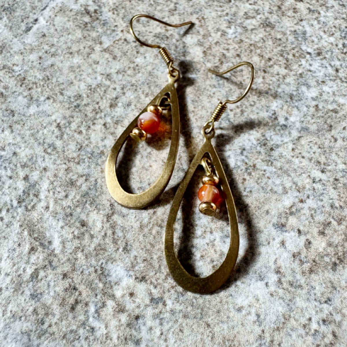 A pair of gold-toned teardrop dangle earrings with small orange and white marbled bead accents, displayed on a textured gray stone surface.