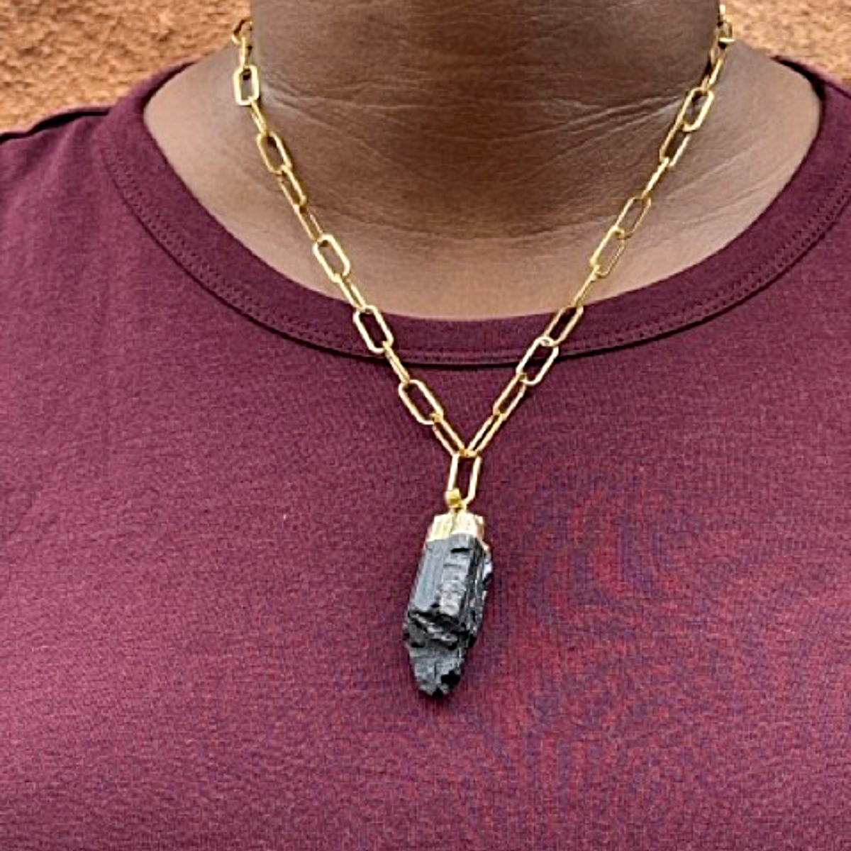 A gold paperclip chain necklace with a raw black gemstone pendant, worn by a person wearing a maroon t-shirt.