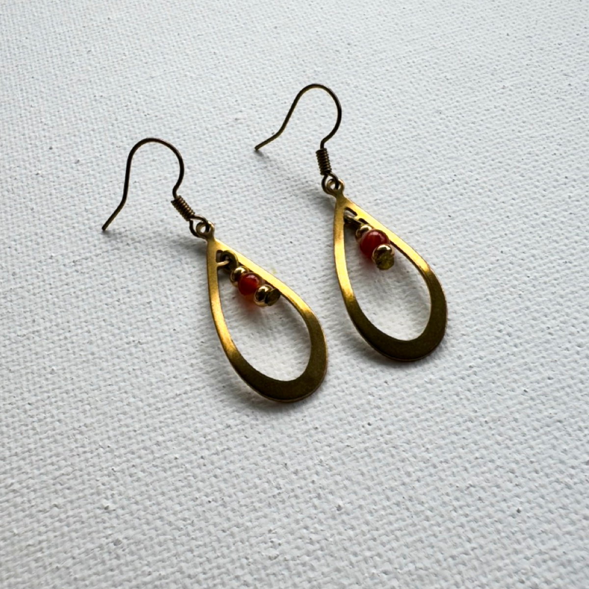 A pair of gold-toned teardrop dangle earrings with small red and gold beads, displayed on a textured white fabric background.