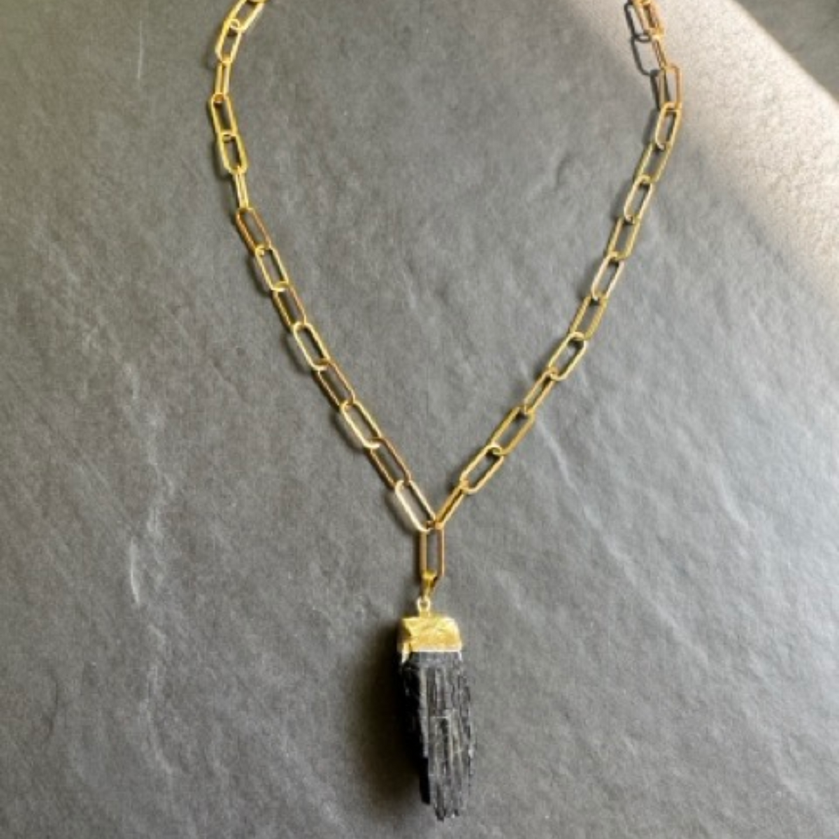 A gold paperclip chain necklace with a  black textured stone pendant, displayed on a textured gray slate surface.