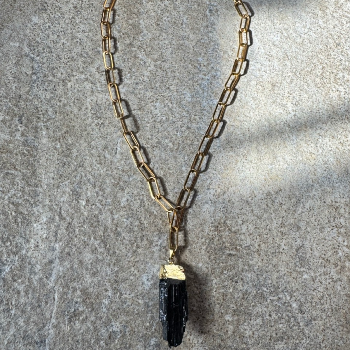 A gold-toned paperclip chain necklace with a dark, textured black stone pendant, laid flat on a textured gray stone surface.