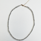 A delicate beaded necklace made of small gray faceted beads with a gold-tone clasp and chain, displayed on a textured white background.