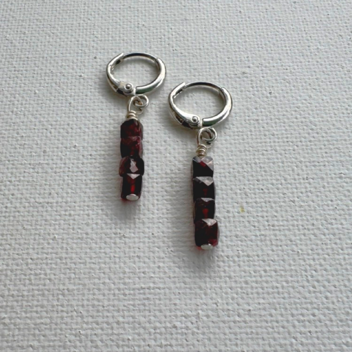A pair of silver hoop earrings with dangling vertical strands of faceted dark red beads, displayed on a textured white fabric background.