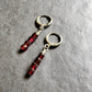 A pair of handmade dangle earrings featuring faceted dark red beads suspended from textured silver-toned metal hoops, displayed on a gray textured surface.