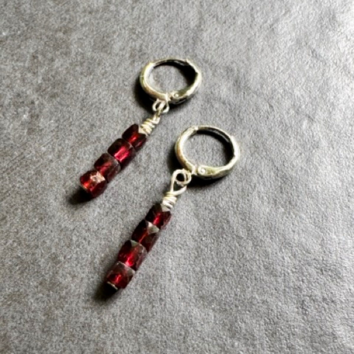 A pair of handmade dangle earrings featuring faceted dark red beads suspended from textured silver-toned metal hoops, displayed on a gray textured surface.