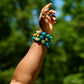 Woman's left arm raised in the air, wearing  a stack of three blue, green, and yellow multicolored bead bracelets, in front of blurred background of green trees.  