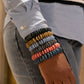 A close-up of a person's wrist wearing a stack of colorful Ashanti bead bracelets, styled with a light blue button-down shirt and blue jeans.