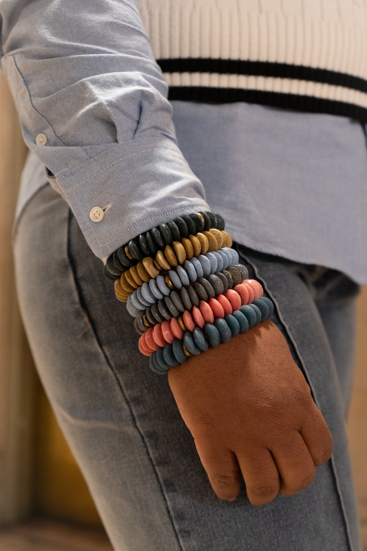 A close-up of a person's wrist wearing a stack of colorful Ashanti bead bracelets, styled with a light blue button-down shirt and blue jeans.