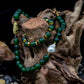 A hunter green beaded necklace with a brass focal bead and a baroque pearl, featuring hematite spacers and lampwork glass beads with matching bracelet on tree trunk against black background.