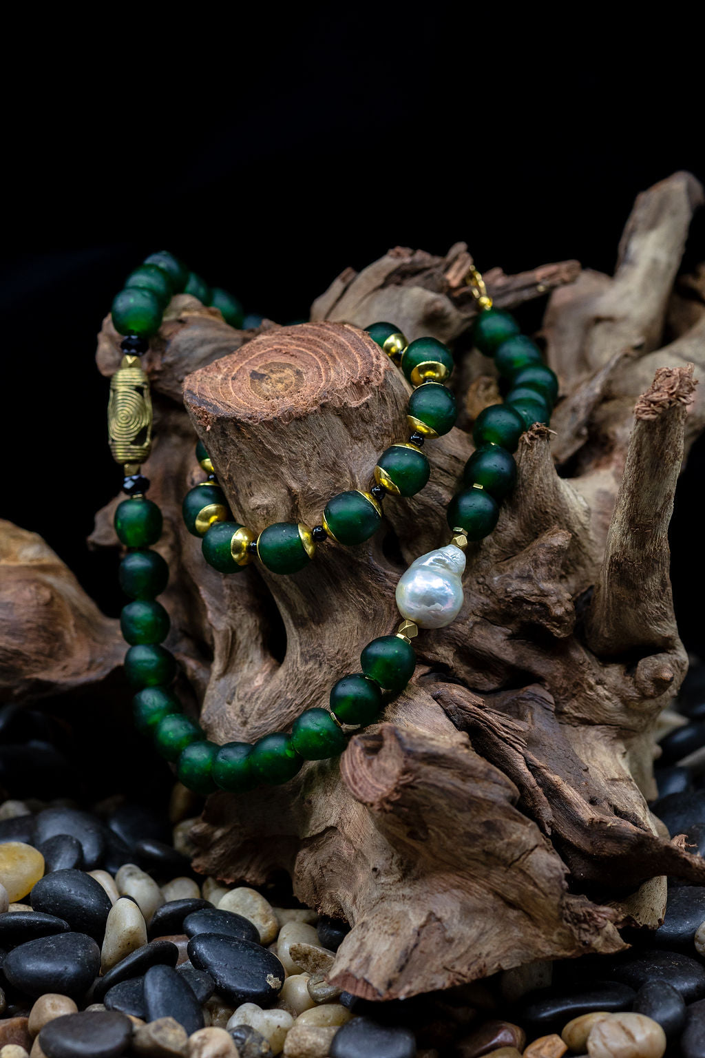 A hunter green beaded necklace with a brass focal bead and a baroque pearl, featuring hematite spacers and lampwork glass beads with matching bracelet on tree trunk against black background.