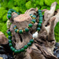 A hunter green beaded necklace with a brass focal bead and a baroque pearl, featuring hematite spacers and lampwork glass beads with matching bracelet on tree trunk against green background.