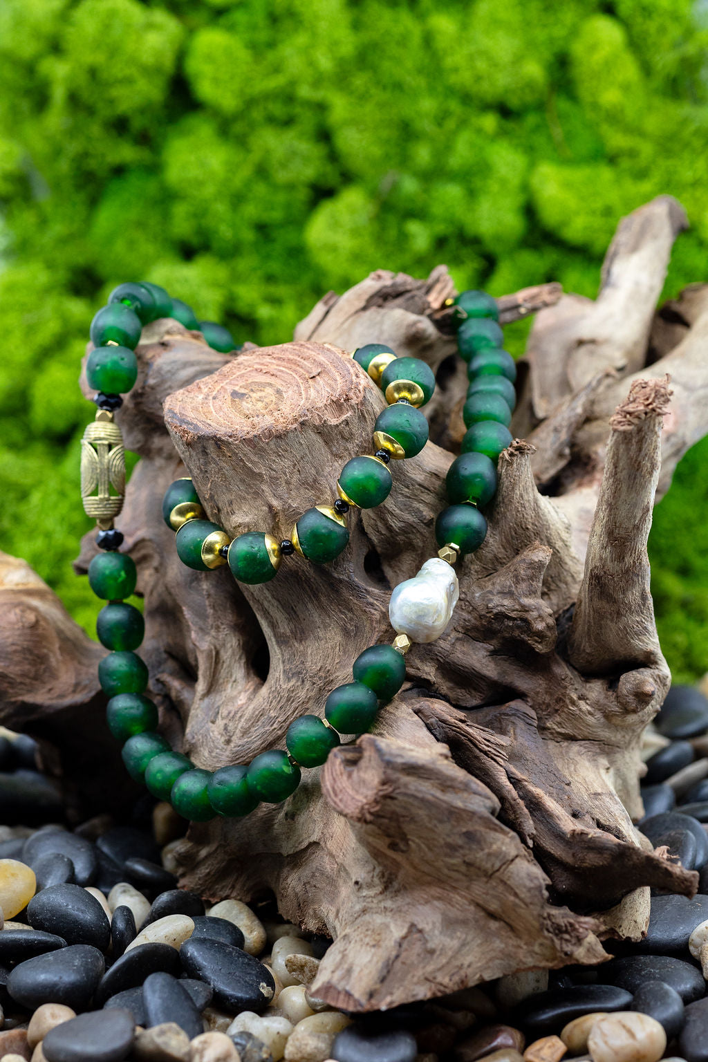 A hunter green beaded necklace with a brass focal bead and a baroque pearl, featuring hematite spacers and lampwork glass beads with matching bracelet on tree trunk against green background.