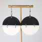Pair of earrings with black and white half-circle design on a gold stand against a light gray background