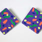 Two purple square earrings with colorful abstract designs on a white background