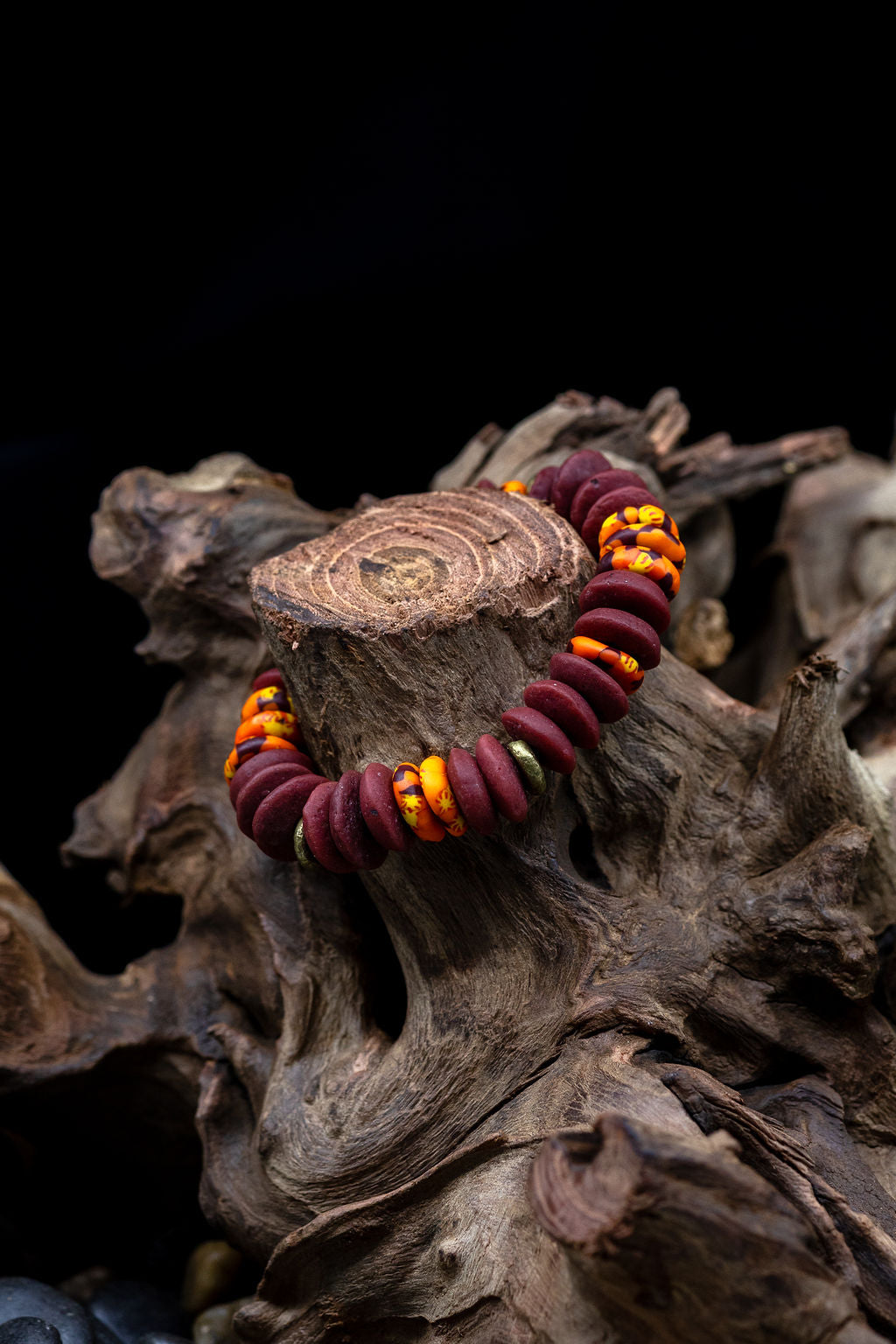 A multicolored bead bracelet with a mix of brown, orange, and yellow hues, featuring brass donut beads and fused rondelle African recycled glass beads on tree trunk against a black background.