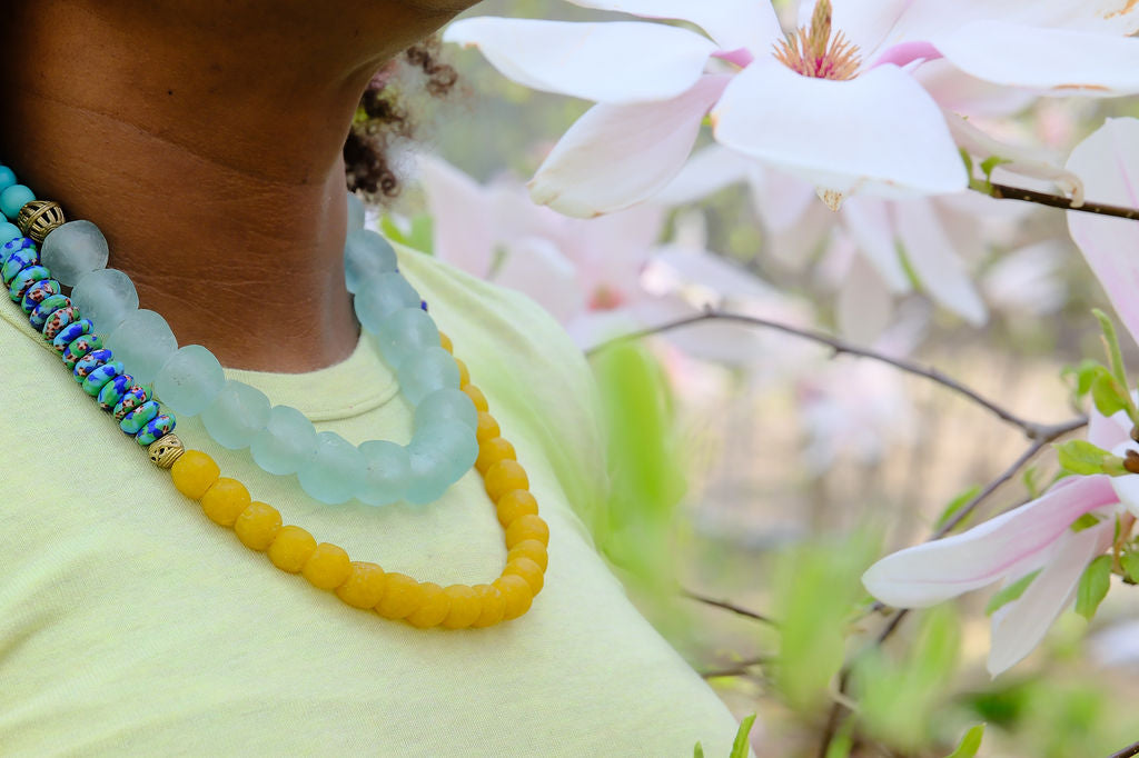 Person wearing colorful beaded necklaces with a blurred background of pink flowers