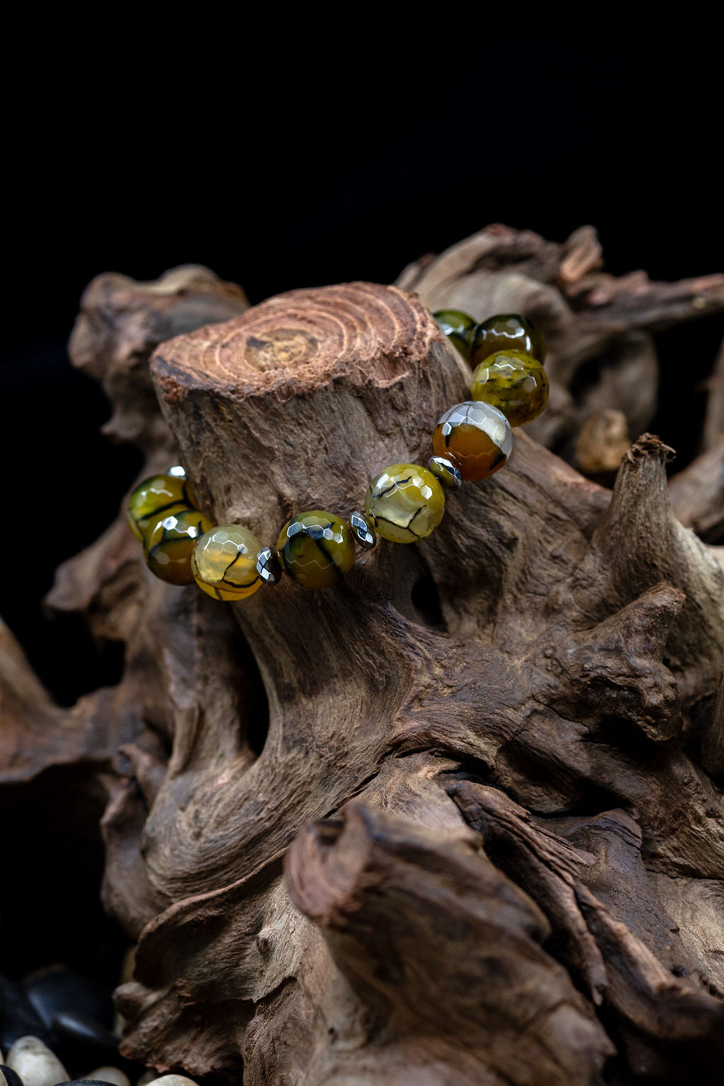 A bead bracelet with a combination of brown, black, and green faceted beads with hematite accents, on a tree stump against a black background. 