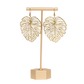 Brass monstera leaf-shaped earrings on a matching stand with a white background