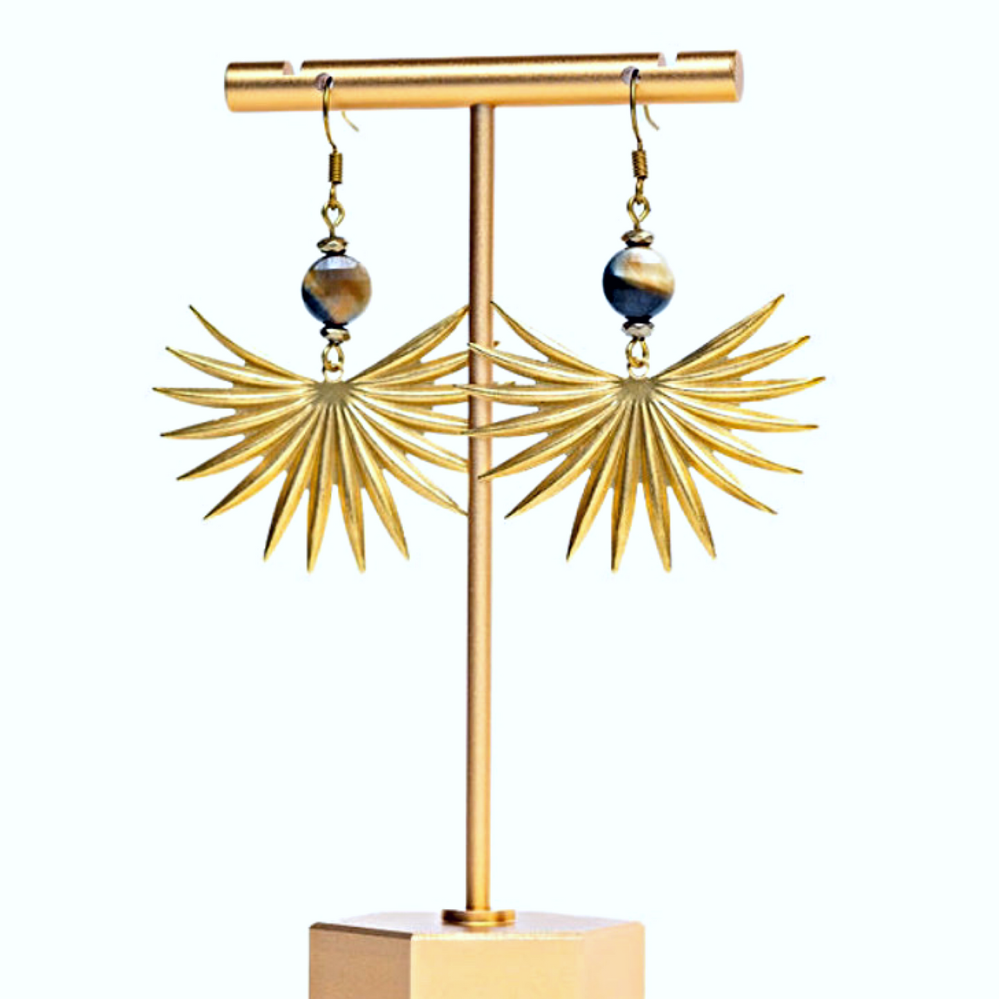 Close up of a pair of earrings featuring brass sun spikes and tiger eye coin beads displayed on a gold-colored stand against white background.