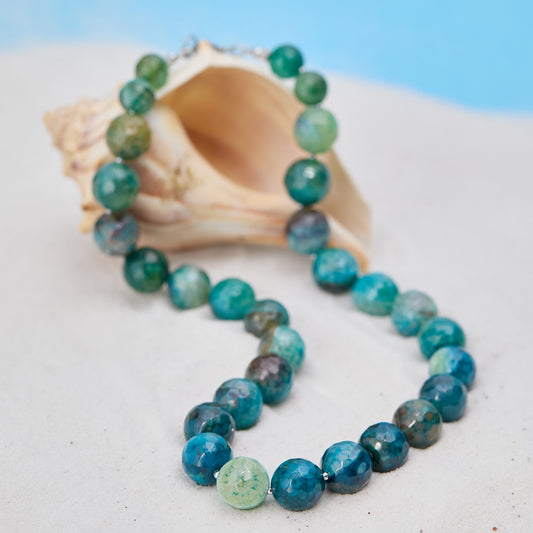 Necklace with blue and green beads on a white surface with a shell in the background